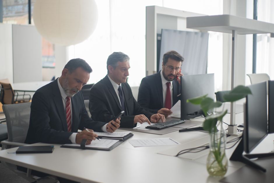 Three men in suits collaborating at a modern office desk with computers and documents.